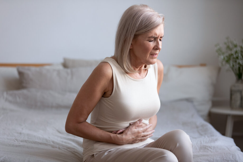 Unhealthy middle aged frowning woman sitting on bed, putting hands on belly, suffering from strong stomach ache.