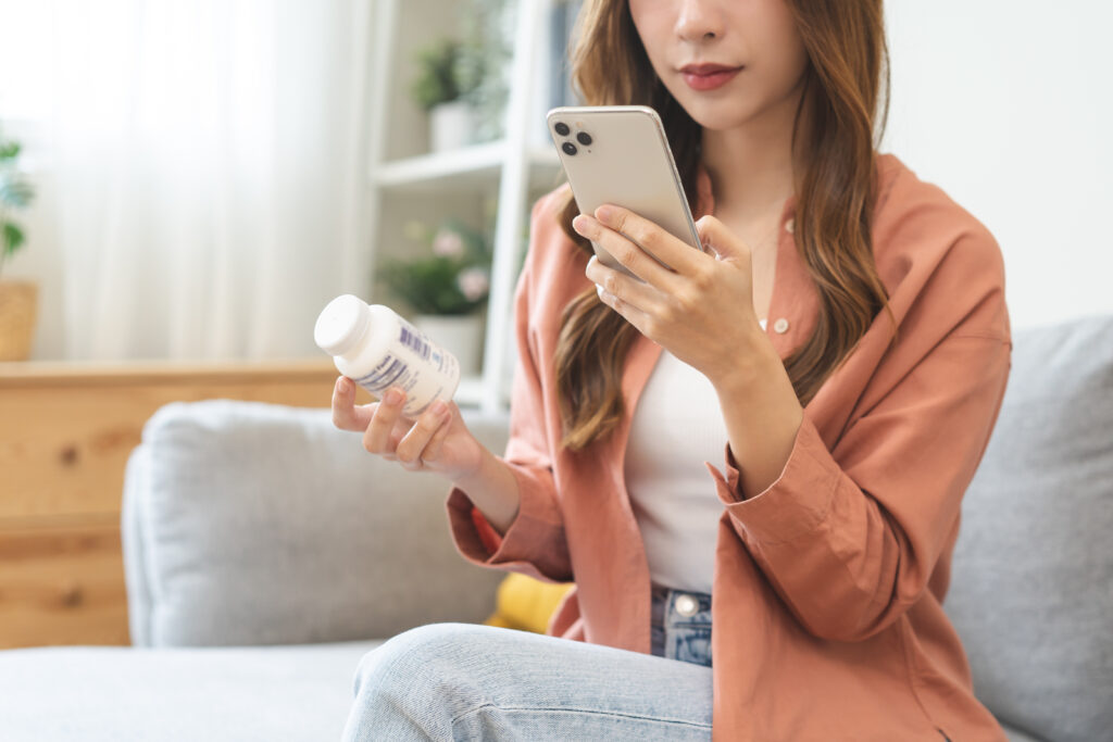 Health care asian young woman using smart phone for reading, searching prescription on bottle medicine, pill label text about information online, instructions side effects, pharmacy medicament concept